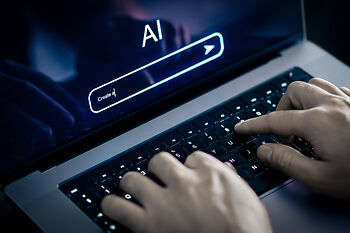 Eine Person tippt auf einer Laptop-Tastatur. Auf dem Bildschirm sind die Wörter 'AI' und ein Texteingabefeld mit der Aufforderung 'Create' zu sehen. Der Hintergrund ist leicht unscharf und dunkel.
