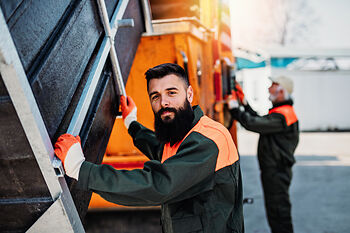 Männer in Arbeitskleidung, einer steht neben einem großen Lkw und schaut in die Kamera, der andere arbeitet im Hintergrund. Beide tragen Handschuhe und sind in einer industrialisierten Umgebung.