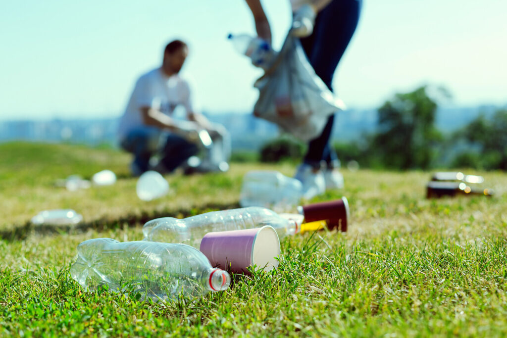 Personen sammeln Abfälle auf einer Wiese, wobei leere Plastikflaschen und Becher auf dem Gras liegen. Im Hintergrund ist eine zweite Person mit einem Müllsack zu sehen.