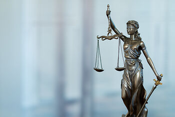 Eine bronzene Statue der Justitia, die eine Waage in der linken Hand hält und ein Schwert in der rechten Hand. Sie trägt eine Augenbinde und eine antike Robe, steht auf einem neutralen verschwommenen Hintergrund.