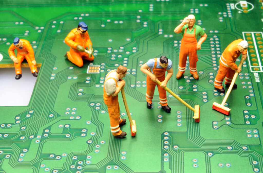 Miniaturfiguren von Arbeitern in orangefarbenen Overalls, die auf einer Leiterplatte mit grüner Farbe und elektronischen Schaltkreisen arbeiten. Einige fegen den Boden, während andere in verschiedenen Positionen stehen.